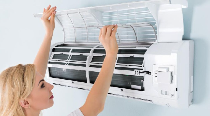 An aerial view of a wastewater treatment facility surrounded by various landscapes, including cultivated fields and a small cluster of residential buildings. The facility features circular water treatment pools and is situated near a road that curves through the area. A hilly terrain extends in the distance with the coastline visible at the horizon.